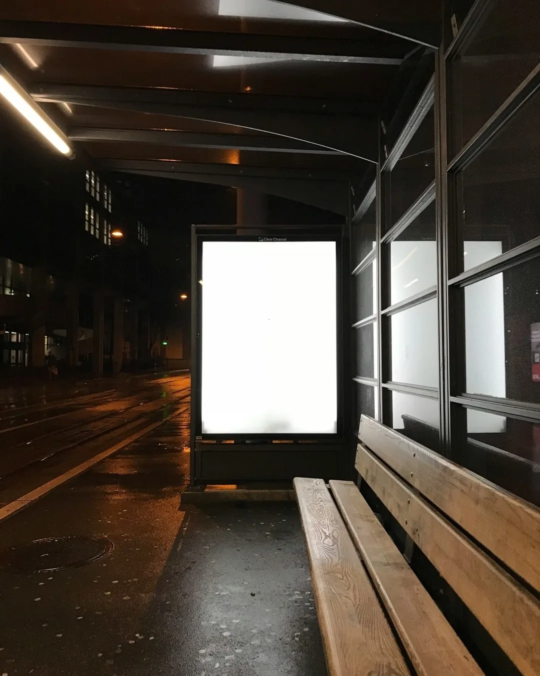 Abri de tramway la nuit sous la pluie. Un caisson publicitaire rétroéclairé occupe le centre du cadre, entièrement blanc et sans affiche. À droite, un banc en bois vide longe une paroi vitrée ; à gauche, une rue mouillée où se reflètent des lampadaires orange.