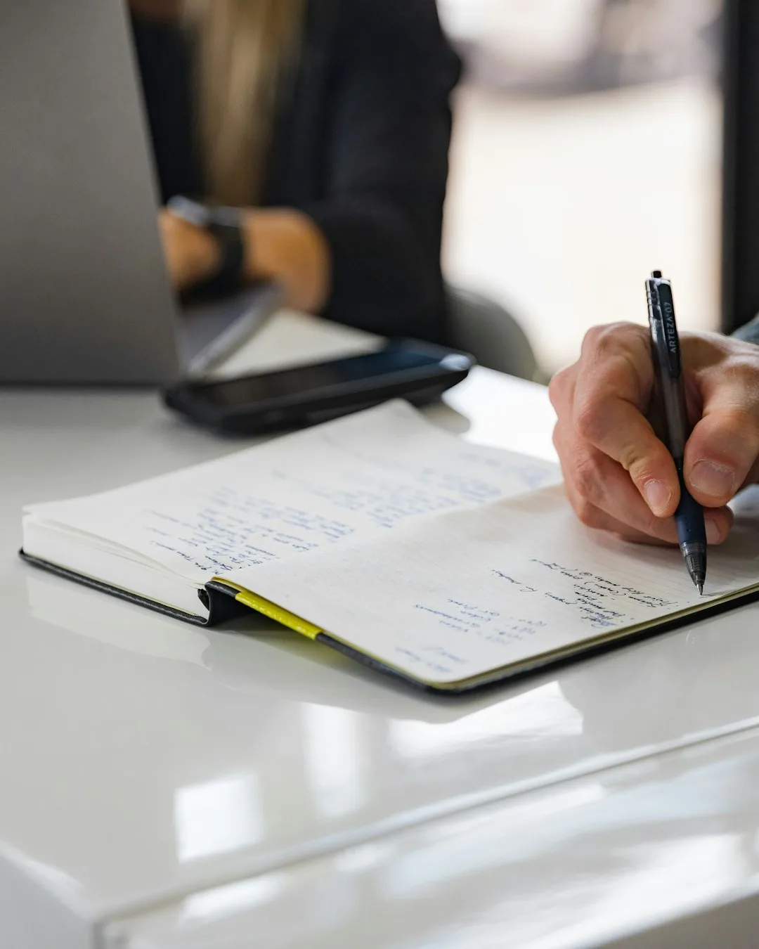 Main prenant des notes au stylo dans un carnet, sur une table de travail, une autre personne travaillant à l'arrière-plan.