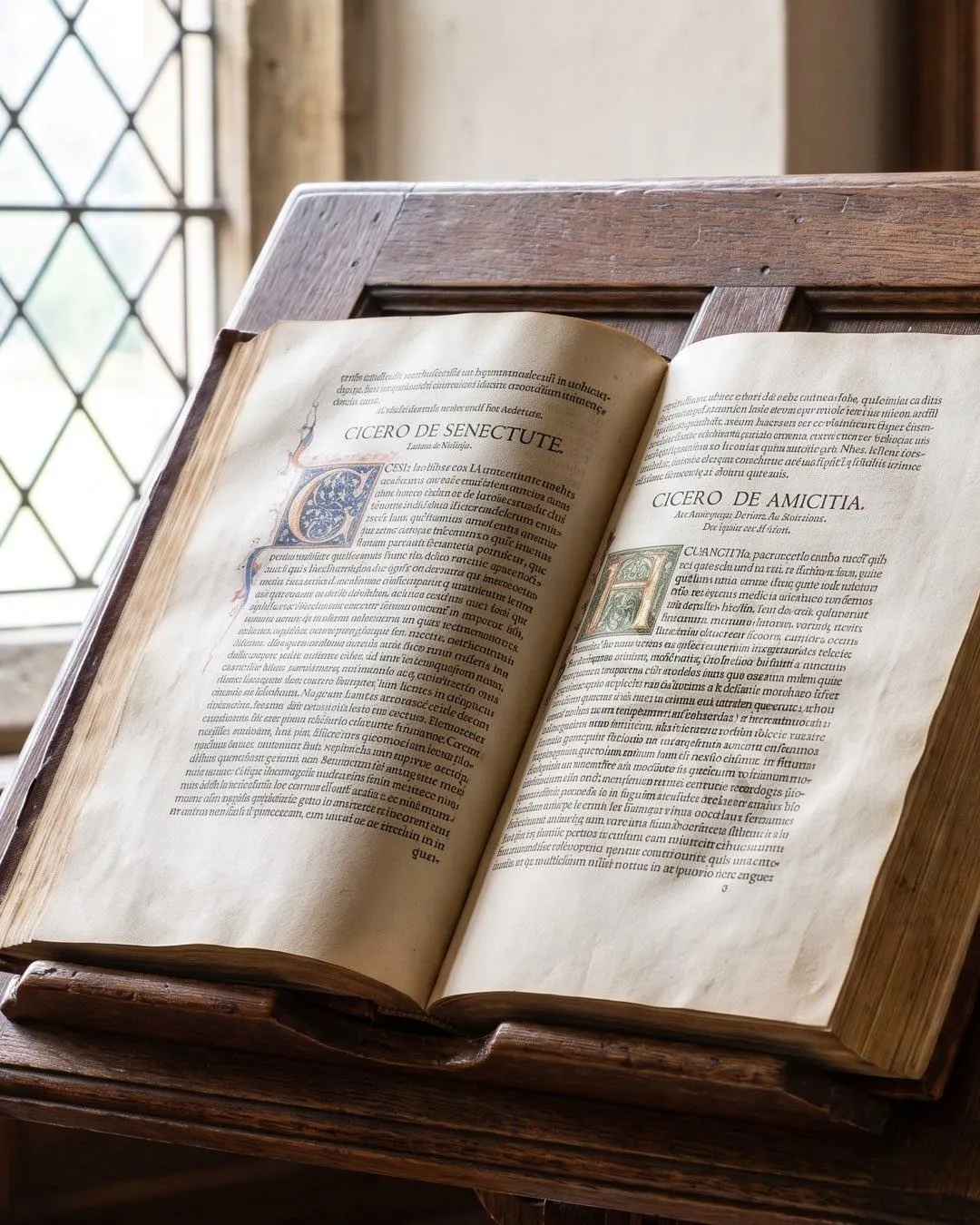 Un livre ancien ouvert sur un lutrin en bois devant une fenêtre à petits carreaux. Deux titres imprimés, Cicero de Senectute et Cicero de Amicitia, avec des lettrines enluminées bleu et or. Le corps du texte est illisible.