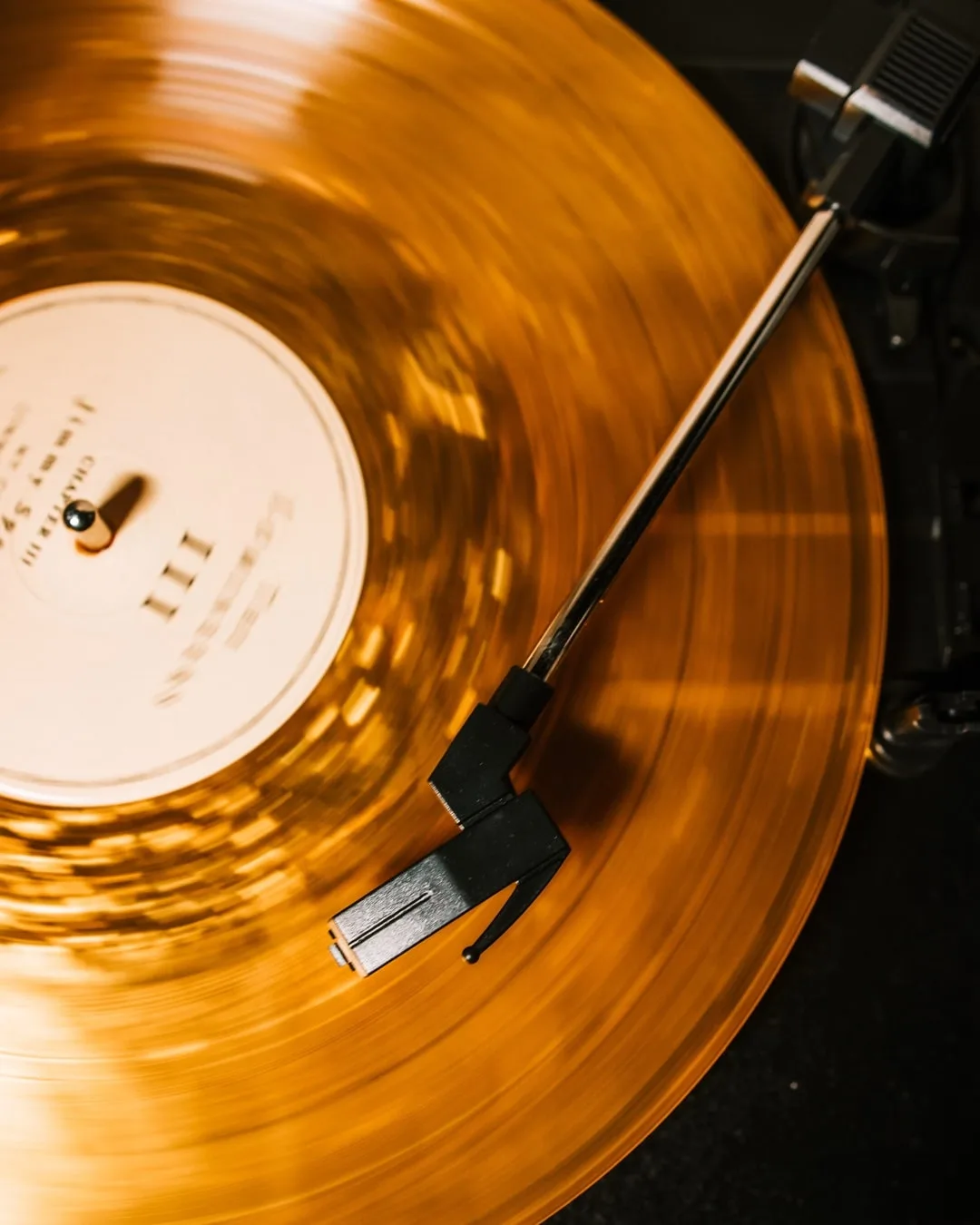 Vue de dessus d'un disque vinyle ambré en rotation sur une platine, le mouvement rendant les sillons flous. Le bras de lecture traverse l'image en diagonale, sa cellule posée dans le sillon.
