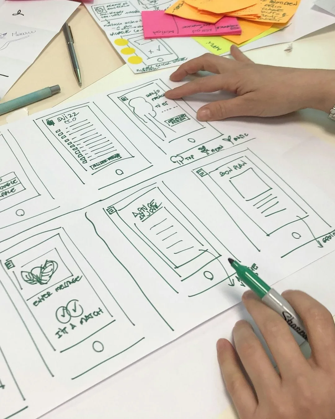 Planche de croquis d'écrans mobiles sur une table d'atelier, deux mains au travail, l'une pointant un écran, l'autre tenant un feutre, post-its en arrière-plan.